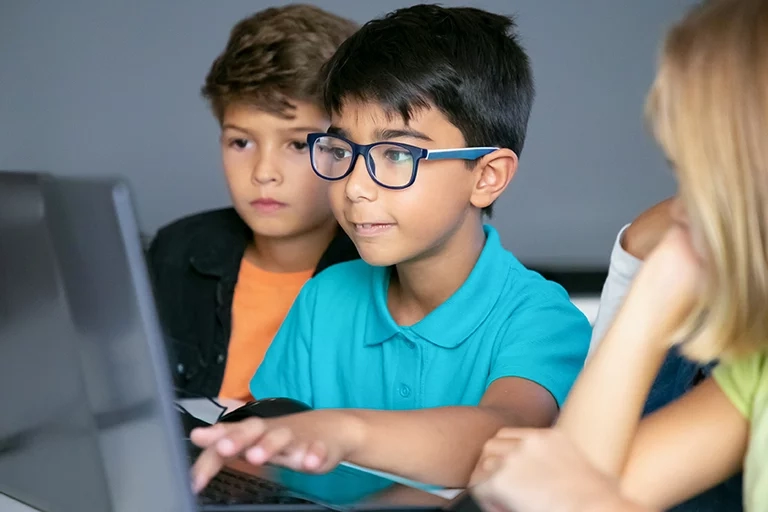 Kid wearing glasses working on a computer with his classmates