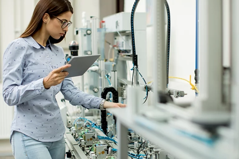 Woman wearing glasses standing next to an assembly line holding an tablet