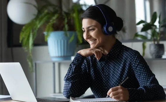 Woman wearing headphones holding a pen over paper smiling at a laptop